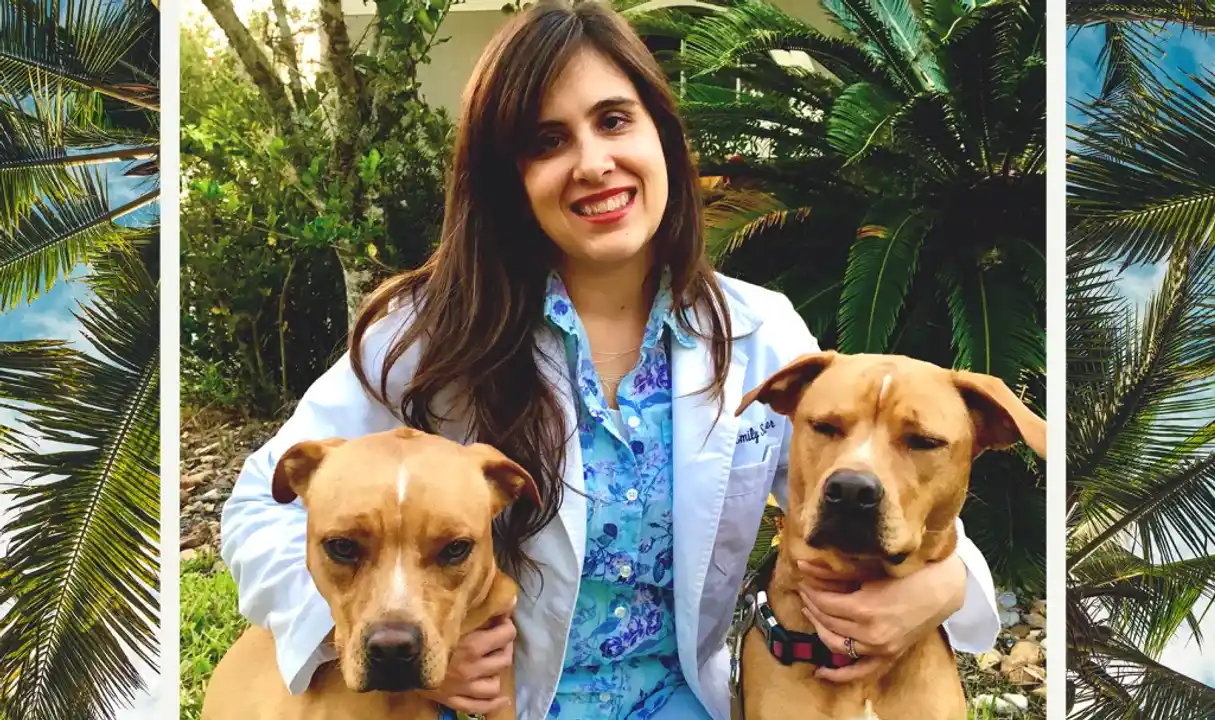 Photograph of a person with long brown hair wearing a lab coat smiles with their arms around two short-haired blonde dogs.