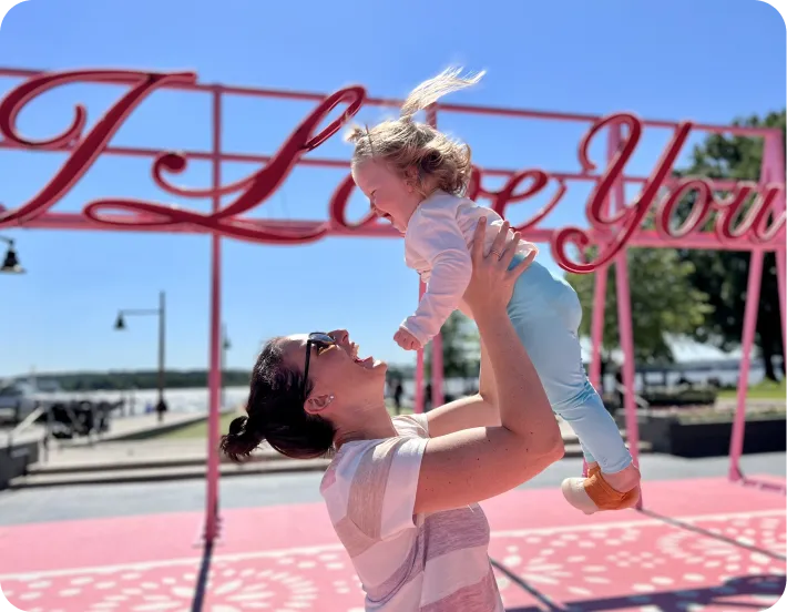 Photo of a mother holding her child in the air in front of a sign reading 