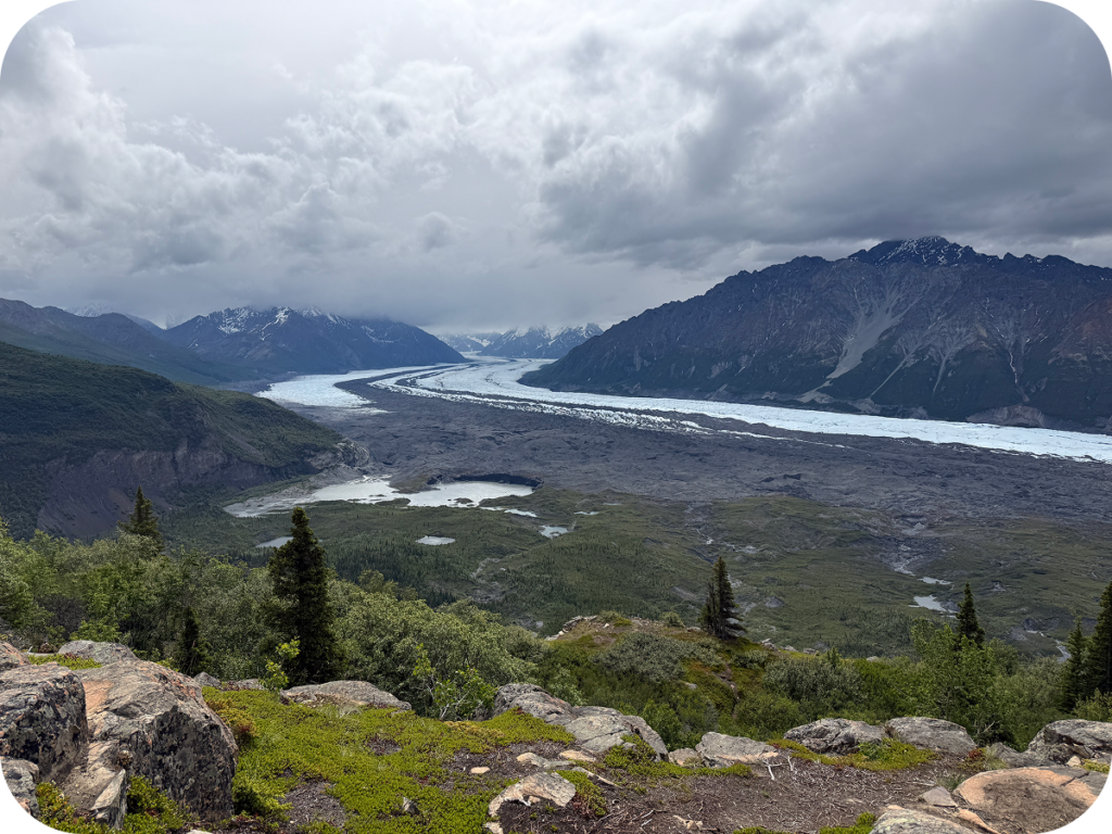 Photo of an Alaskan landscape with snow-covered stretches of land weaving between stout mountains beneath a grey, cloudy sky